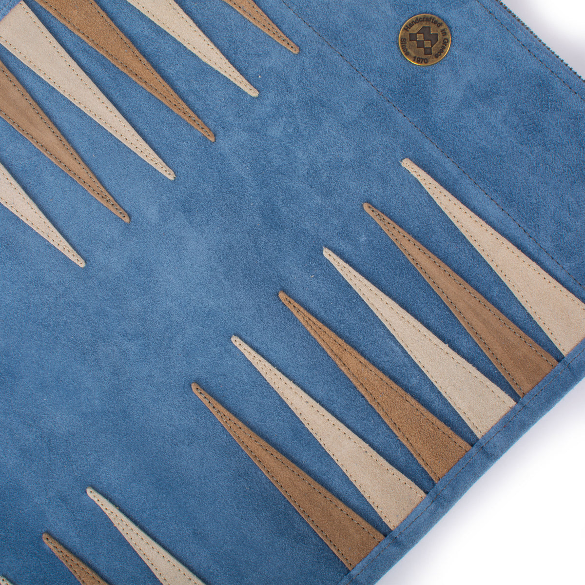 Close-up of a denim blue suede backgammon board with tan and cream stitched points and a circular Manopoulos emblem, showcasing luxury Greek craftsmanship