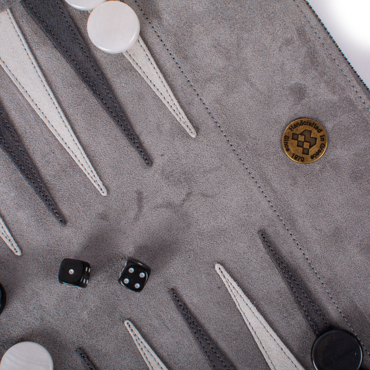 Close up of a grey suede roll up backgammon board with stitched black and white points, black dice and a Manopoulos emblem, showcasing handcrafted Greek design