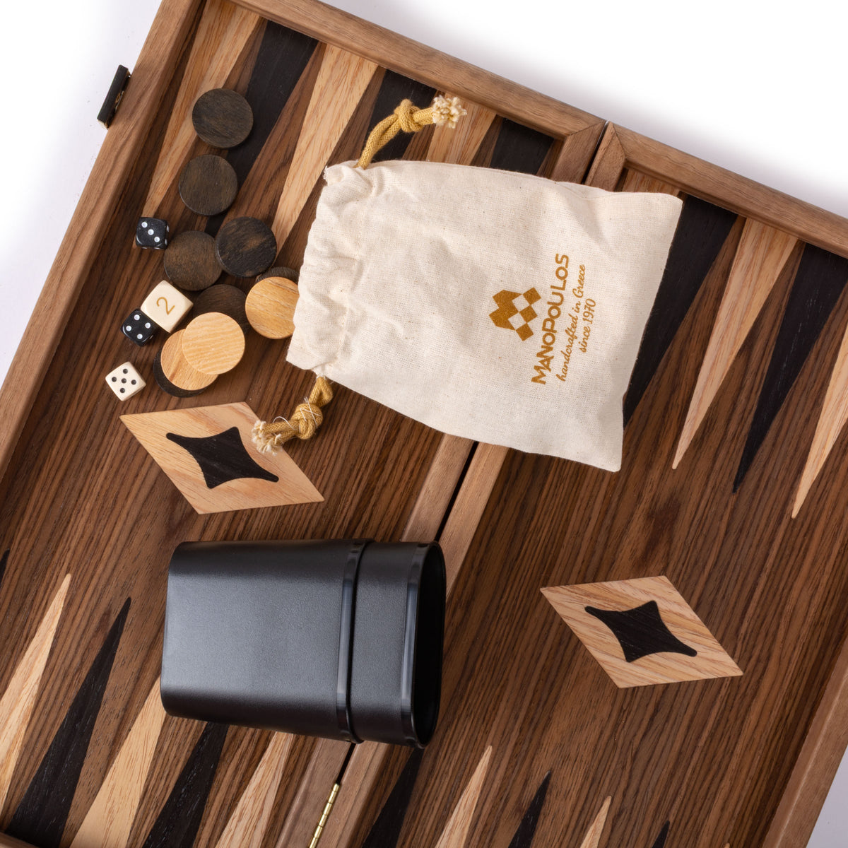 Open wooden backgammon board with dark and light striped inlay, checkers, dice, leather dice cup, and drawstring pouch arranged on the playing surface.